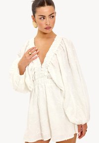 Witte geborduurde jurk met pofmouwen, diepe V-hals, gerimpelde taille en bloemenpatronen, gemaakt van een textuurrijke, lichte stof.