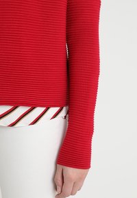 Rechter Arm in einem gerippten roten Langarmoberteil über weißen Hosen, mit einem gestreiften Hemdenstück, das am Bund herausragt.