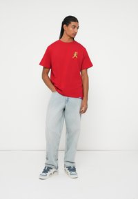 Rød bomulds T-shirt med gul grafisk print på brystet, parret med lyseblå løse jeans og grå sneakers.