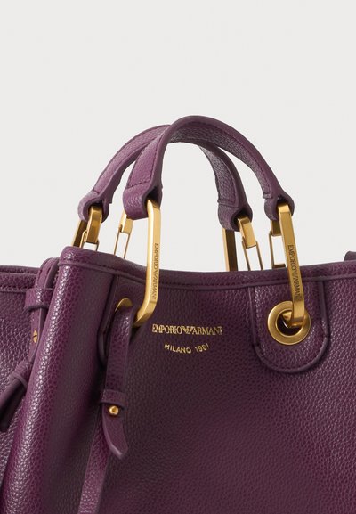 Μωβ υφασμάτινη δερμάτινη τσάντα Emporio Armani με χρυσούς λαβές και λογότυπο σφραγισμένο με "Milano 1981" σε χρυσά γράμματα.