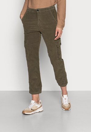 Personne portant un pantalon cargo vert olive avec poches latérales, un pull beige et des baskets blanc-beige, debout sur un sol gris devant un mur blanc.