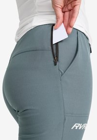 Graue Sporthosen mit Rippstruktur, die eine Reißverschlusstasche auf der Rückseite haben. Eine Hand steckt eine Karte in die Tasche.