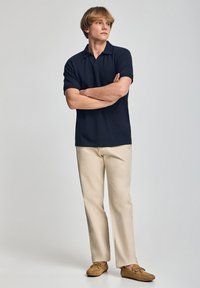 Camisa polo de color azul marino oscuro con mangas cortas, combinada con pantalones beiges y zapatos mocasín marrones. Diseño sencillo, colores sólidos y corte casual.