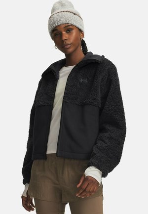 Vrouw draagt een grijze gebreide muts, een zwarte getextureerde fleece jas, een wit shirt met lange mouwen en bruine gelaagde shorts over legging.