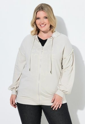 Smilende kvinde iført en lysebeige hættejakke med lynlås, snører og puffede ærmer, kombineret med mørke jeans, mod en hvid baggrund.