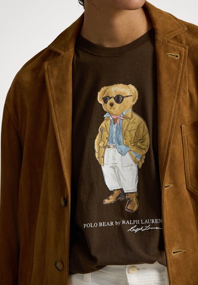 Brązowa zamszowa marynarka na brązowej koszulce z nadrukiem kreskówkowego misia w beżowej kurtce, niebieskiej koszuli i okularach przeciwsłonecznych, noszącego białe spodnie i buty.