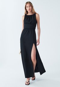 Schwarzes Maxikleid mit ärmellosem Design, gerafftem Ausschnitt, geraffter Taille, Seitenschlitz und glatter Textur. Getragen mit schwarzen Flip-Flops.