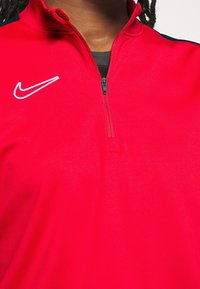 Punainen Nike-puolihihainen pusero, jossa on sileä pinta, valkoinen logo vasemmassa rinnassa ja tummansiniset yksityiskohdat harteilla.