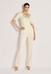 Vrouw poseert in een lichtgele top met korte mouwen en bloemenrand, gecombineerd met crème brede broek en gouden hoge hakken.