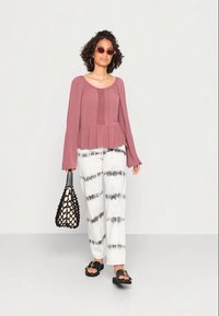 Top peplum em rosa envelhecido com mangas longas, combinado com calças brancas apresentando padrões de tie-dye em preto. Acessorizado com uma bolsa tecida preta.