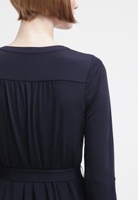 Marinblå långärmad klänning med figursydd bodice, samlade detaljer bak och knytning i midjan. Slät tygkvalitet med subtil glans och rundad halsringning.