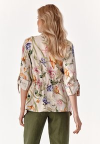 Bluse i let, blomstrede linned med indsnøret talje, trekvartærmer med opslået manchet og en blød beige baggrund.