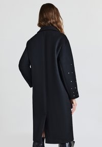 Lungo cappotto nero realizzato in tessuto liscio, con uno spacco sul retro e maniche decorate con piccoli chiodini decorativi.