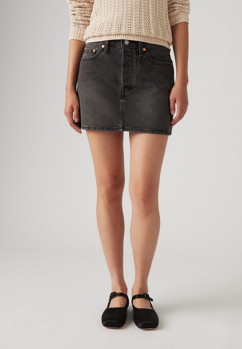 Jupe en denim noir avec une coupe ajustée, courte, cinq poches, et des coutures visibles. Associée à des chaussures Mary Jane noires.