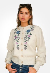 Joven mujer con cabello castaño claro lleva un cárdigan de punto crema con bordado floral azul y morado y jeans de cintura alta azules.