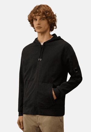 Giovane uomo con capelli ricci che indossa una felpa nera con zip e tasche e pantaloni beige, in piedi con una mano in tasca.