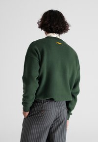 Zielony sweter z dzianiny z prążkowanym okrągłym dekoltem i opuszczonymi ramionami; charakteryzuje się subtelnym, żółtym logo. W połączeniu z spodniami w prążki.