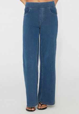 Weit geschnittene blaue Denim-Jeans mit elastischem Bund, getragen mit schwarzen Flip-Flops, Vorderansicht von der Taille bis zu den Füßen.
