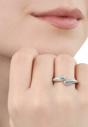 Nahaufnahme einer Hand mit einem glatten, offenen silbernen Ring am Mittelfinger, nahe den Lippen mit einem leichten Lächeln.