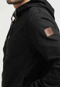 Czarna kurtka z kapturem zapinana na zamek. Posiada brązową skórzaną łatę na lewym rękawie oraz gładką, matową fakturę.