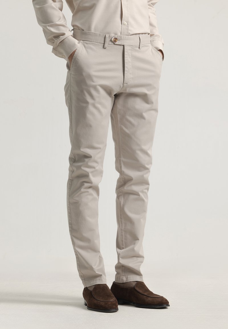 Pantalones de vestir de algodón beige con un diseño ajustado, dos bolsillos laterales, cierre de botón y una textura sutil, combinados con zapatos oscuros de ante.