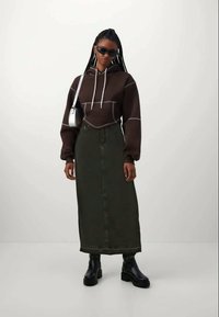 Ruskea huppari, jossa on valkoiset narut, pitkä oliivinvihreä farkkuhame, mustat korkeat saappaat ja aurinkolasit. Sileä kangas ja jämäkkä istuvuus.