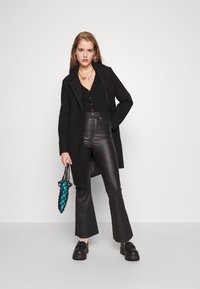 Casaco longo preto sobre um top preto justo com botões, combinado com calças de pele pretas de cintura alta e cortadas em flare. Segura uma bolsa texturizada turquesa.