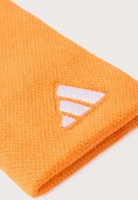 Pomarańczowa tkanina o teksturowanym wykończeniu, z białym haftowanym logo przedstawiającym trzy nakładające się na siebie trójkąty na krawędzi.