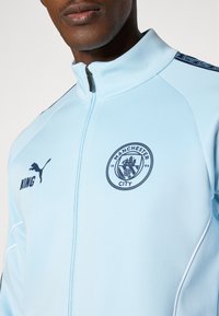 Light blue jacket made of smooth fabric, featuring a standing collar, zip closure, and Manchester City logo in navy. Puma logo on the left.