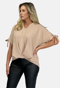 Beżowa, luźna bluzka z dekoltem w kształcie litery V i węzłem z przodu, z podwiniętymi rękawami z troczkami. W zestawie z czarnymi spodniami.
