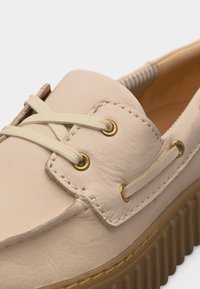 Mocassin en cuir beige avec semelle en caoutchouc épaisse et rainurée, lacets beiges passés à travers des œillets dorés et couture détaillée.