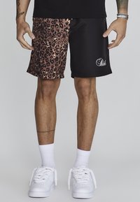 Pantaloni corti con stampa leopardata nera e marrone, con una finitura liscia, caratterizzati da un lato nero con logo bianco. Indossati con sneaker bianche e calze.