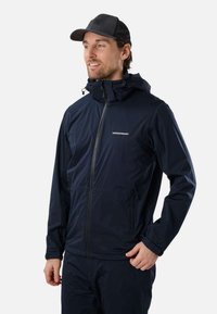 Homme portant une veste zippée bleu marine avec le logo "Swedemount" et une casquette noire, regardant sur le côté avec un léger sourire.