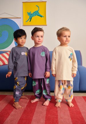 Trīs basām kājām staigājoši mazuļi, kas ģērbti krāsainās pidžamās ar kosmosa tēmu, stāv uz sarkana svītraina paklāja dzīvojamā istabā ar zilu dīvānu un abstraktu sienas mākslu.