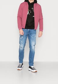 Rosa Bomberjacke über einem schwarzen Hoodie, kombiniert mit zerrissenen blauen Jeans und schwarz-weißen Sneakers. Legeres Outfit mit verschiedenen Texturen.