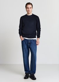 Marineblauer Strickpullover mit Rundhalsausschnitt, blaue Jeans mit geradem Bein und schwarze Lederschuhe, auf einem neutralen Hintergrund arrangiert.
