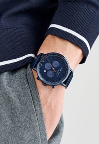 Reloj cronógrafo de color navy con tres subesferas, caja metálica y correa texturizada. Lleva puesto en una muñeca con una prenda gris de fondo.