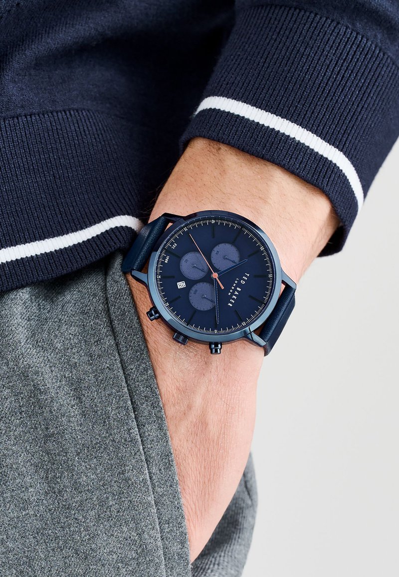 Reloj cronógrafo de color navy con tres subesferas, caja metálica y correa texturizada. Lleva puesto en una muñeca con una prenda gris de fondo.