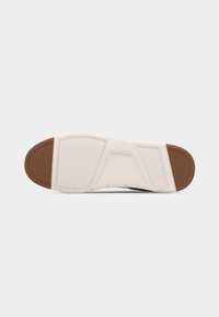 Η σόλα ενός αθλητικού παπουτσιού με λευκή βάση και καφέ ελαστικά τμήματα στη φτέρνα και τα δάχτυλα, με υφασμένο μοτίβο πρόσφυσης σε επίπεδη επιφάνεια.