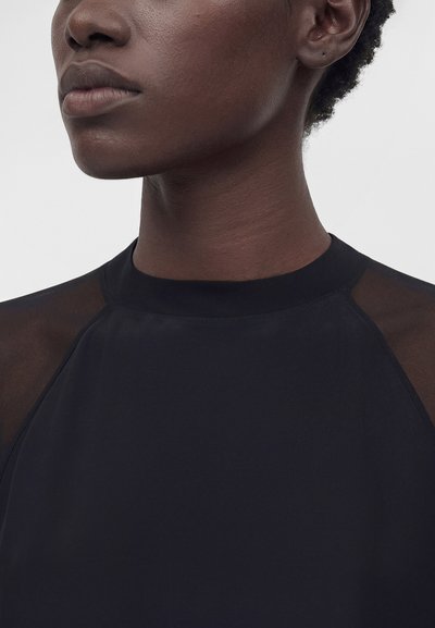 Κοντινό πλάνο ενός ατόμου με σκουρόχρωμο δέρμα που φοράει μαύρο τοπ με διαφανείς πήχεων ώμων από δίχτυ, εμφανιζόμενο από τον λαιμό έως το πάνω μέρος του στήθους.