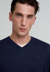 Pulover bleumarin cu guler în V, fabricat dintr-un material tricotat moale, având un mic logo cu craniu pe piept și un tricou bej stratificat pe dedesubt.