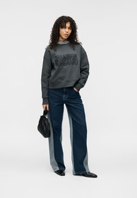 Γκρι φούτερ με έντονο μαύρο κείμενο, συνδυασμένο με φαρδιά μπλε τζιν με μοτίβο κοψίματος. Το μοντέλο κρατά μια μαύρη τσάντα με υφή.