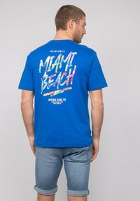 T-shirt en coton bleu avec un graphique "MIAMI BEACH" aux couleurs vives dans le dos, avec une coupe décontractée et des manches courtes. Associé à un short en denim.