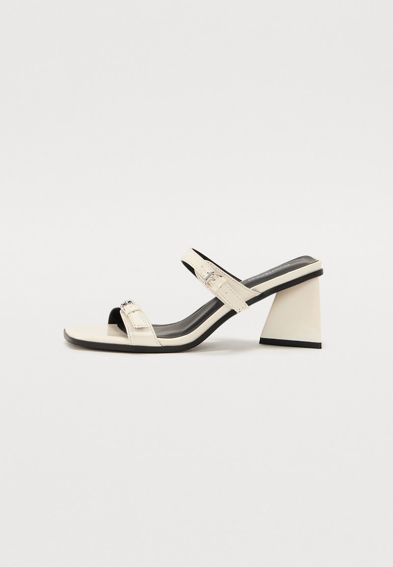 Sandalias de tacón en color blanco roto con dos tiras finas, puntera cuadrada y un tacón geométrico y bloque. Acabado en cuero suave con acentos en negro.