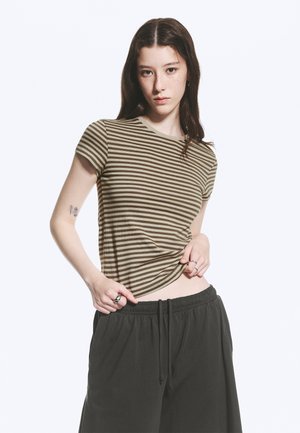 Junge Frau mit langen dunklen Haaren trägt ein braun-beigefarben gestreiftes T-Shirt und schwarze, lockere Hosen, hebt ihr Shirt leicht mit einer Hand an.