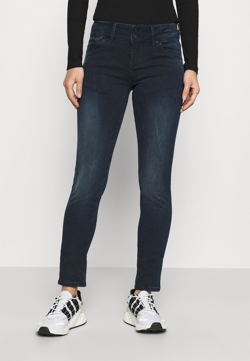 Femme portant un jean slim bleu foncé avec des baskets noires et blanches sur un fond clair uni.