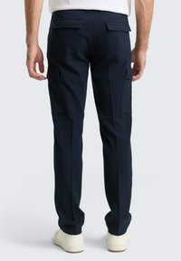 Mann trägt navyblaue Cargo-Hosen mit Klappentaschen auf der Rückseite und den Seiten, kombiniert mit weißen Turnschuhen und einem weißen Hemd.