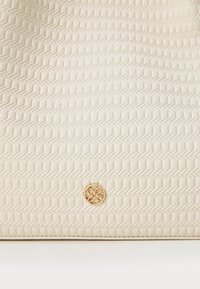 Biała skórzana torebka z fakturowanym wzorem w zygzak oraz złotym, okrągłym logo umieszczonym na środku u dołu.