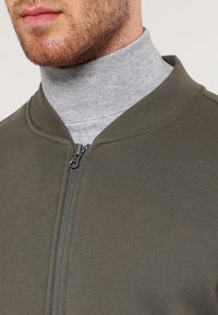 Olive green zip-up jacket with a ribbed collar and cuffs, worn over a light gray turtleneck, featuring a smooth texture and minimal hardware.