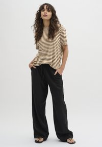 Chemise à manches courtes rayée beige et blanche associée à un pantalon large noir. Le modèle porte des tongs noires ; coupe décontractée, tissu doux.